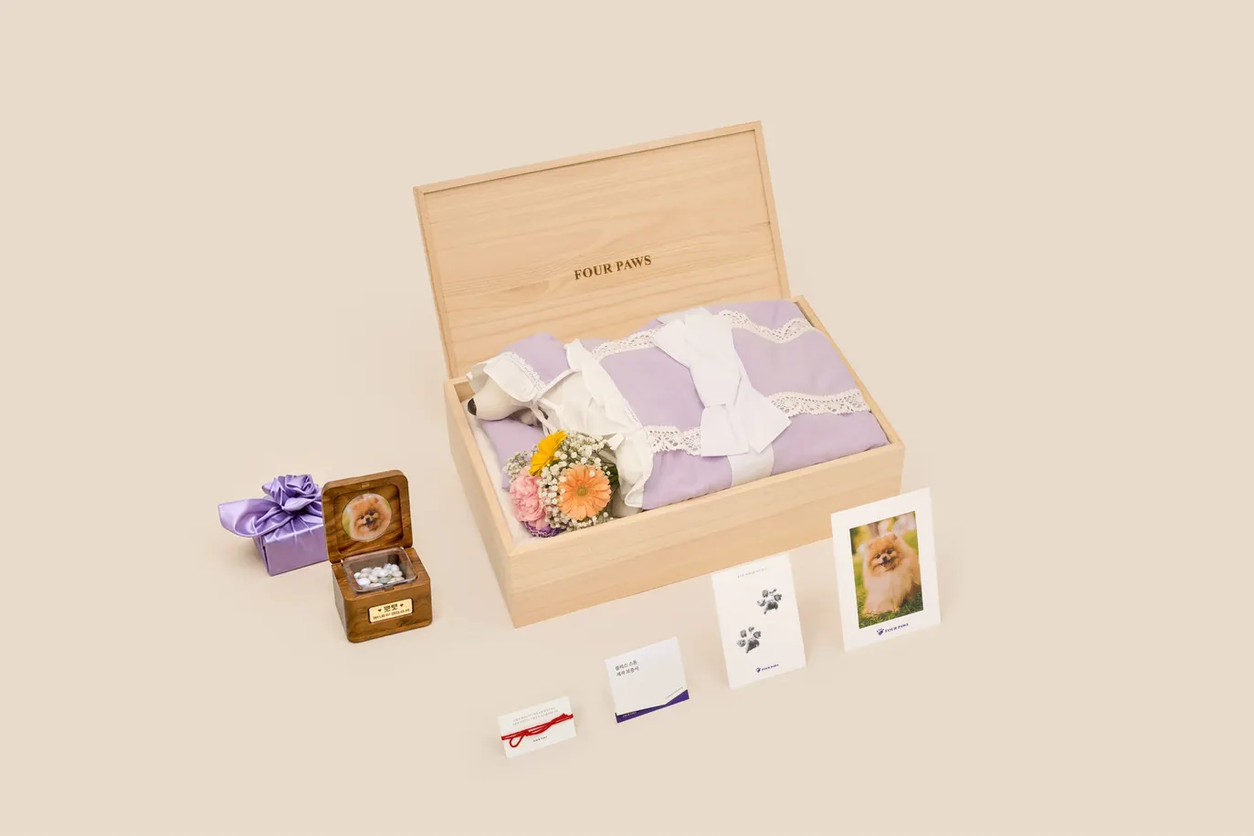 연 오동나무 관 안에 연보라 수의를 입은 강아지 모형과 꽃다발이 누워 있고, 관 왼편에 뚜껑 안쪽에 포메라니안 사진을 넣고 투명 창 너머로 흰 알갱이가 보이는 호두나무 함과 보라색 보자기 상자가 놓인 구성