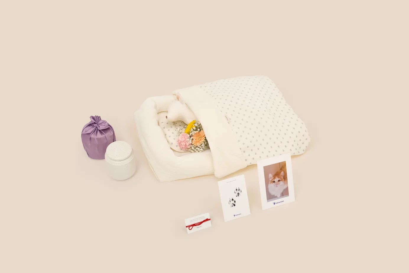 화성 고양이 장례식장에서 쓰는 고양이 전용 장례 구성 — 흰 누빔 이불 모양의 일체형 관에 작은 동물 모형이 누워 있고 그 곁에 분홍·노랑 꽃다발, 보라색 보자기에 싸인 유골함과 흰 도자기 유골함, 발도장 카드와 고양이 사진 카드, 붉은 실이 놓인 모습