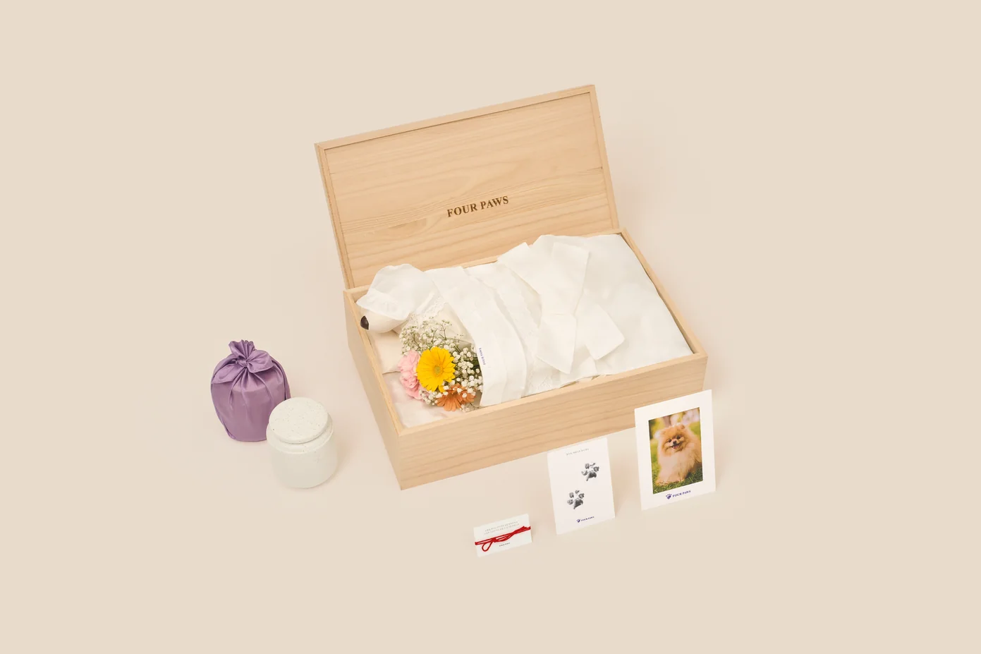 연한 나뭇결의 관 안에 흰 수의를 입은 강아지 모형과 작은 꽃다발이 누워 있고, 관 왼편에 보라색 보자기 꾸러미와 점무늬 뚜껑의 흰 도자기 함, 앞쪽에 인연의 실 카드·발도장 카드·포메라니안 사진 액자가 놓인 구성