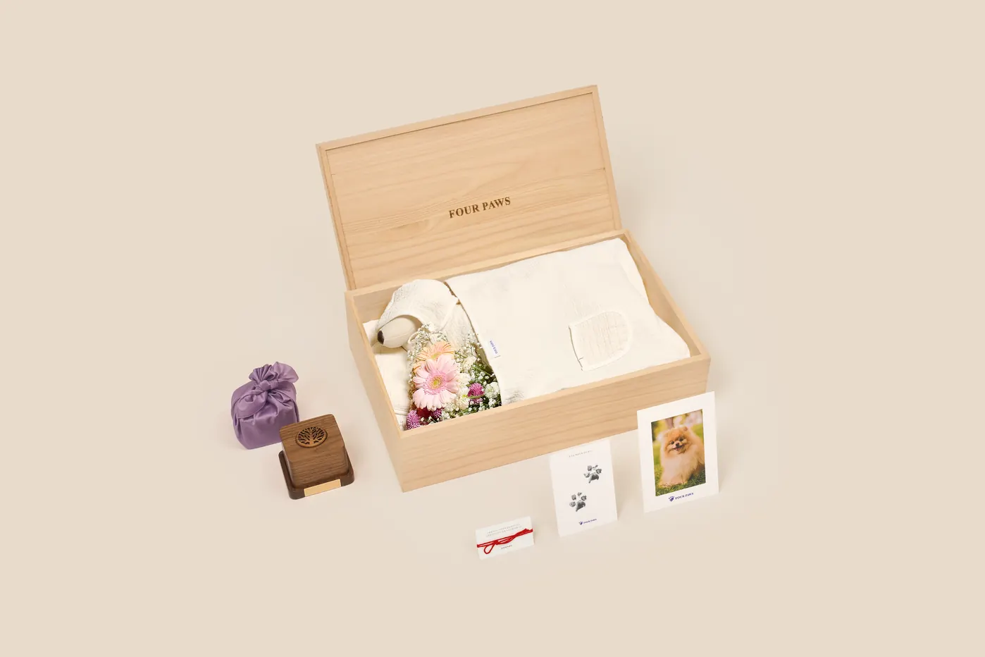 뚜껑을 연 밝은 원목 관 안에 흰 거즈 수의와 분홍 거베라 꽃다발이 담기고, 앞에 보라색 보자기 꾸러미와 짙은 원목 함, 발도장 카드와 빨간 실 카드, 강아지 사진 액자가 놓인 관 장례 구성
