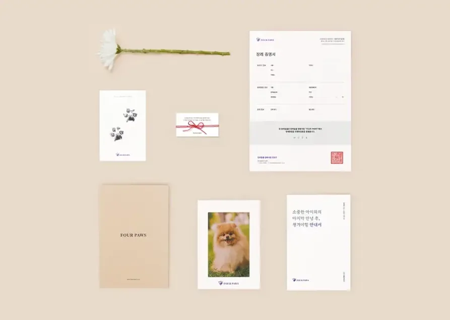 베이지색 바탕에 흰 데이지 한 송이, 발도장 카드, 붉은 실로 묶은 작은 카드, 장례 증명서, FOUR PAWS 글씨의 갈색 봉투, 포메라니안 사진, 장례 후 안내서가 가지런히 놓인 추모 키트