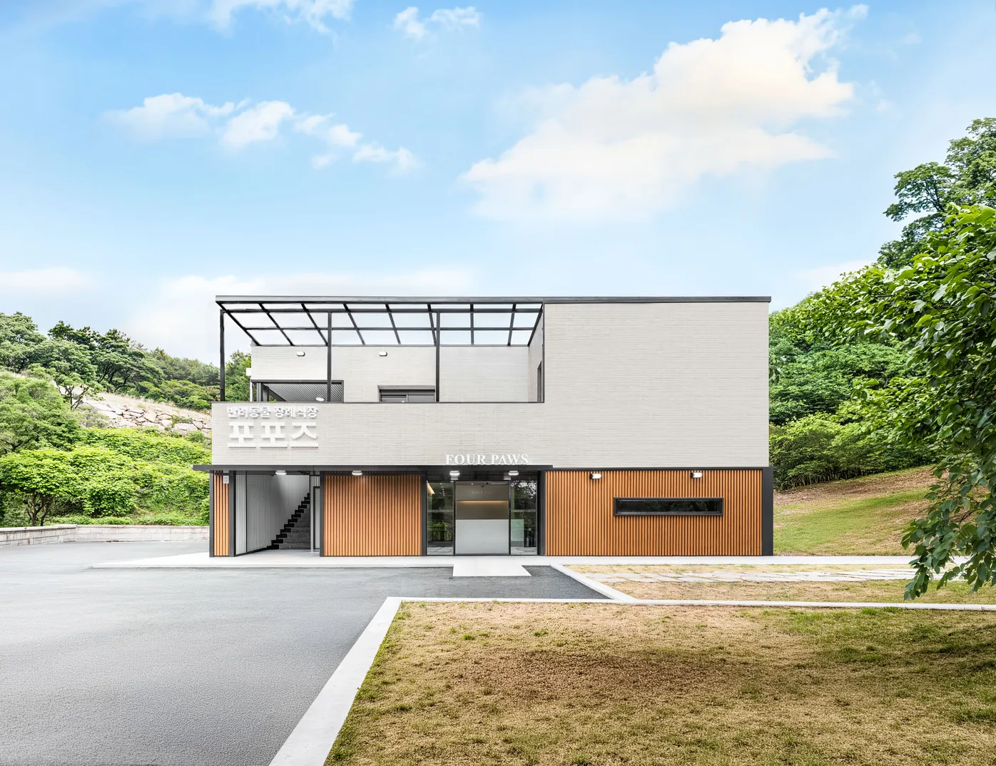 숲을 등지고 선 포포즈 부산점 건물 — 흰 벽돌 벽과 나무 루버 외벽, 앞마당의 잔디와 주차장