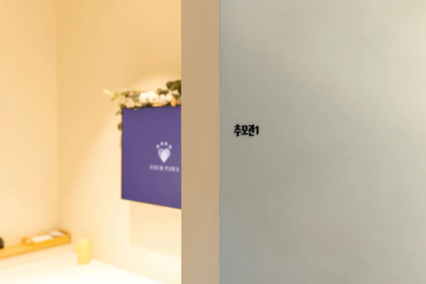「추모관1」 표지가 붙은 문 너머로 보랏빛 판과 꽃 장식이 보이는 포포즈 김포점 추모실