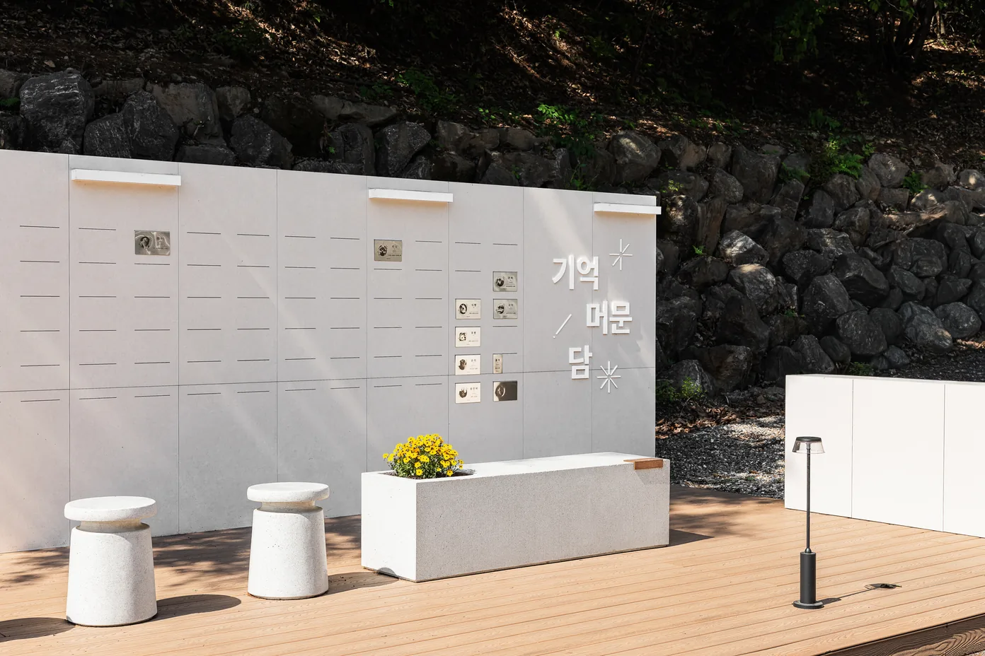경기 광주점 기억 머문 담 — 「기억 머문 담」 글자가 붙은 흰 벽에 작은 명패들이 걸려 있고, 앞에 노란 꽃 화분을 올린 돌 벤치와 둥근 돌 의자 두 개, 뒤로 돌담과 나무가 보이는 야외