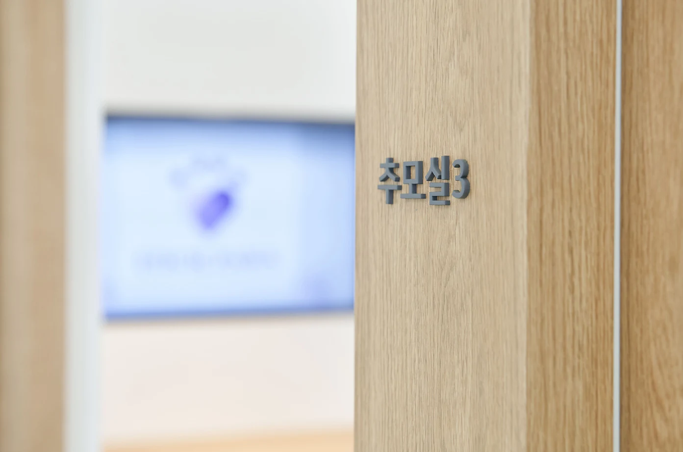 나뭇결 문틀에 추모실3 글자가 붙어 있고 안쪽으로 화면이 흐릿하게 보이는 화성 1호점 추모실 입구